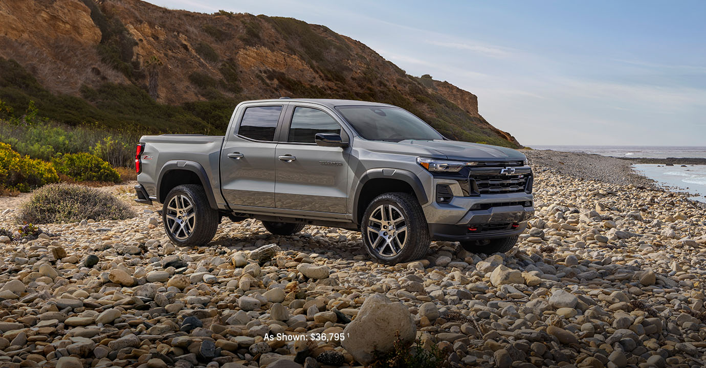 A silver 2025 Chevrolet Colorado parked on a rocky beach with cliffs and the ocean in the background