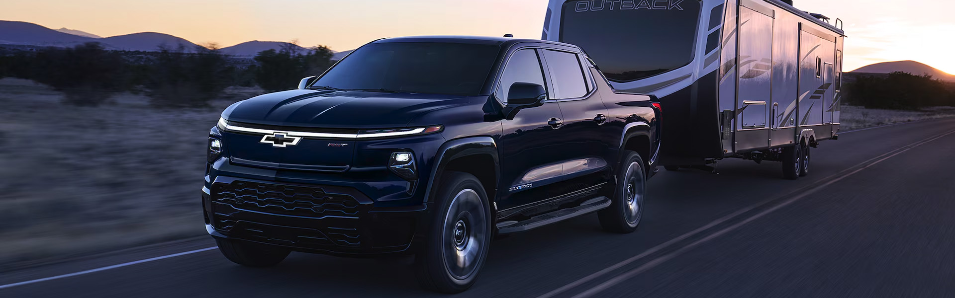 2025 Chevrolet Silverado EV pickup truck towing a large silver travel trailer on an open highway at sunset.