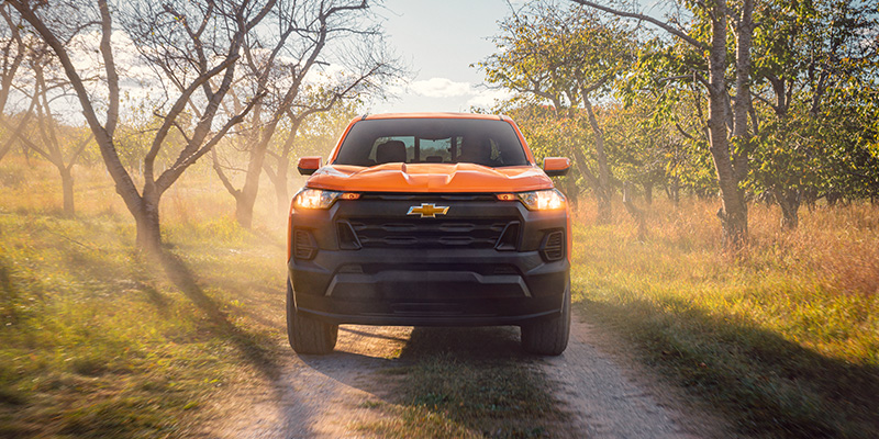 Orange Colorado truck drives down a dusty rural path surrounded by trees under a clear sky, conveying a sense of adventure and freedom.
